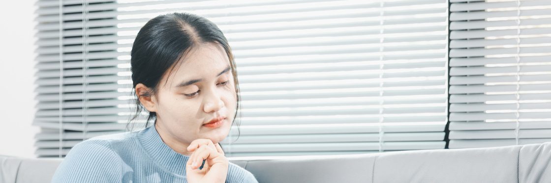 A person sitting on a sofa in front of a window blind, one hand raised near their chin, looking down and thinking.