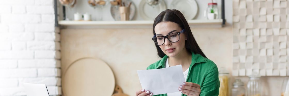 A woman standing in a kitchen, holding up a single printed letter in both hands and reading it.